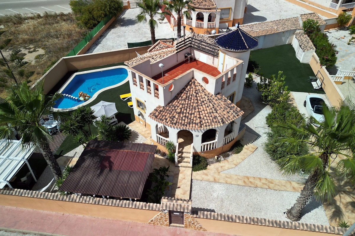 An aerial view of a spacious villa showcases a multi-level structure with a distinctive tiled roof. The private pool is visible alongside a well-maintained yard featuring palm trees and various outdoor seating areas. A parking area is also included in the landscape.