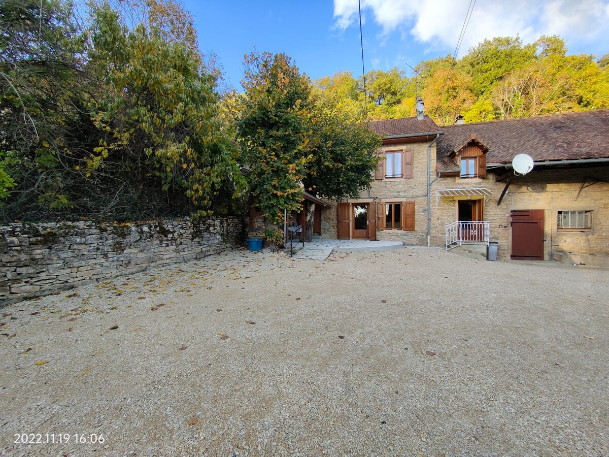 A gravel courtyard is surrounded by stone walls and vegetation, providing a spacious area in front of the rustic farmhouse. The building features light brown wooden shutters and a large entryway, with trees displaying autumn foliage in the background.
