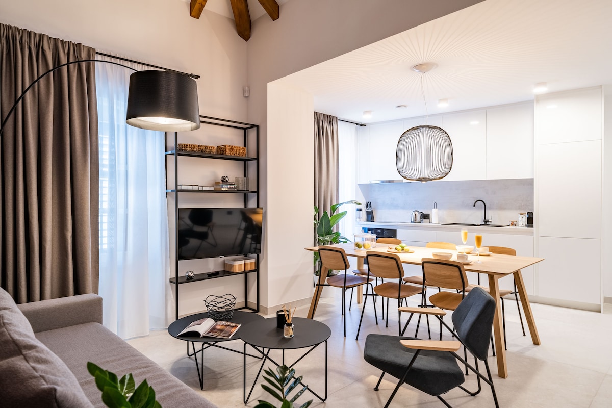 An open-concept living space features modern furnishings, including a cozy grey sofa and sleek dining table. Large windows are adorned with light and dark curtains, allowing natural light to fill the room. Decorative plants add a touch of greenery to the space.