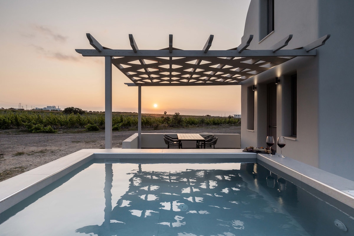 A private patio features a swimming pool reflecting the sunset. A wooden dining table and chairs are positioned beneath a trellis. Glasses of wine and a small platter are placed on the ledge, while gentle vineyards are visible in the background.