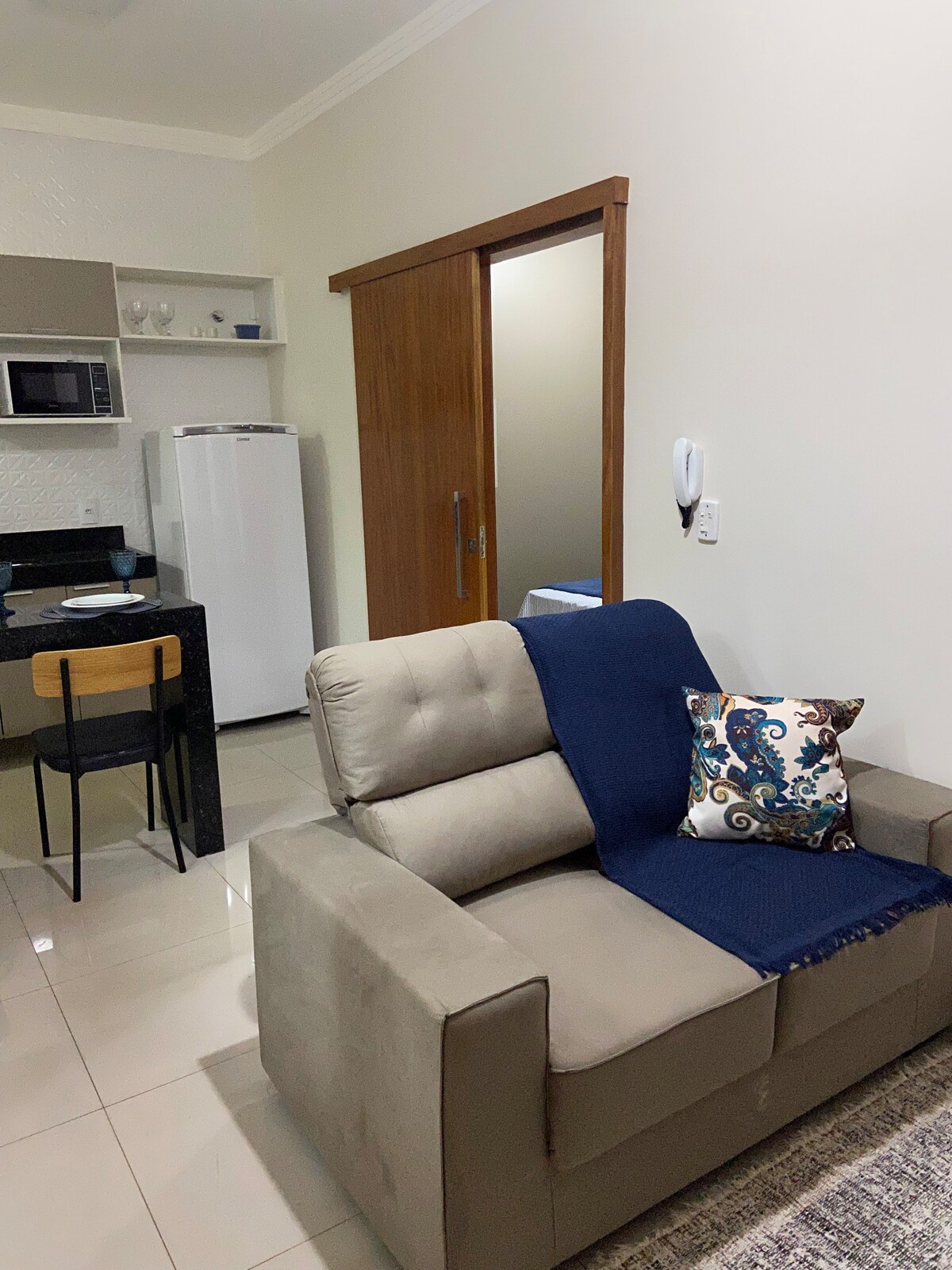A cozy seating area features a soft, neutral-toned armchair draped with a blue throw blanket. A decorative pillow adds a pop of color. In the background, a kitchenette is visible, equipped with a microwave and a refrigerator, while a wooden door leads to another room.