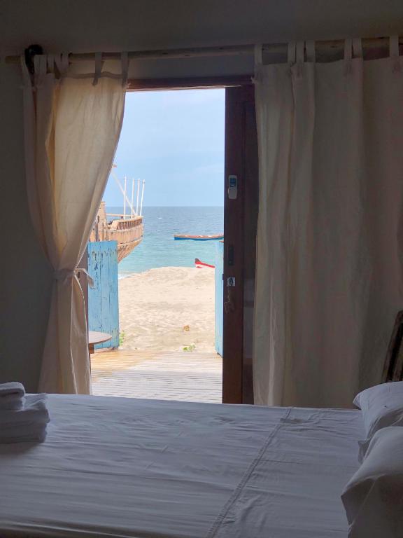 The image captures a serene view from a room, featuring a wooden door that opens to a sandy beach and the ocean. Soft curtains frame the entrance, allowing natural light to illuminate the interior space, with a neatly made bed visible in the foreground.