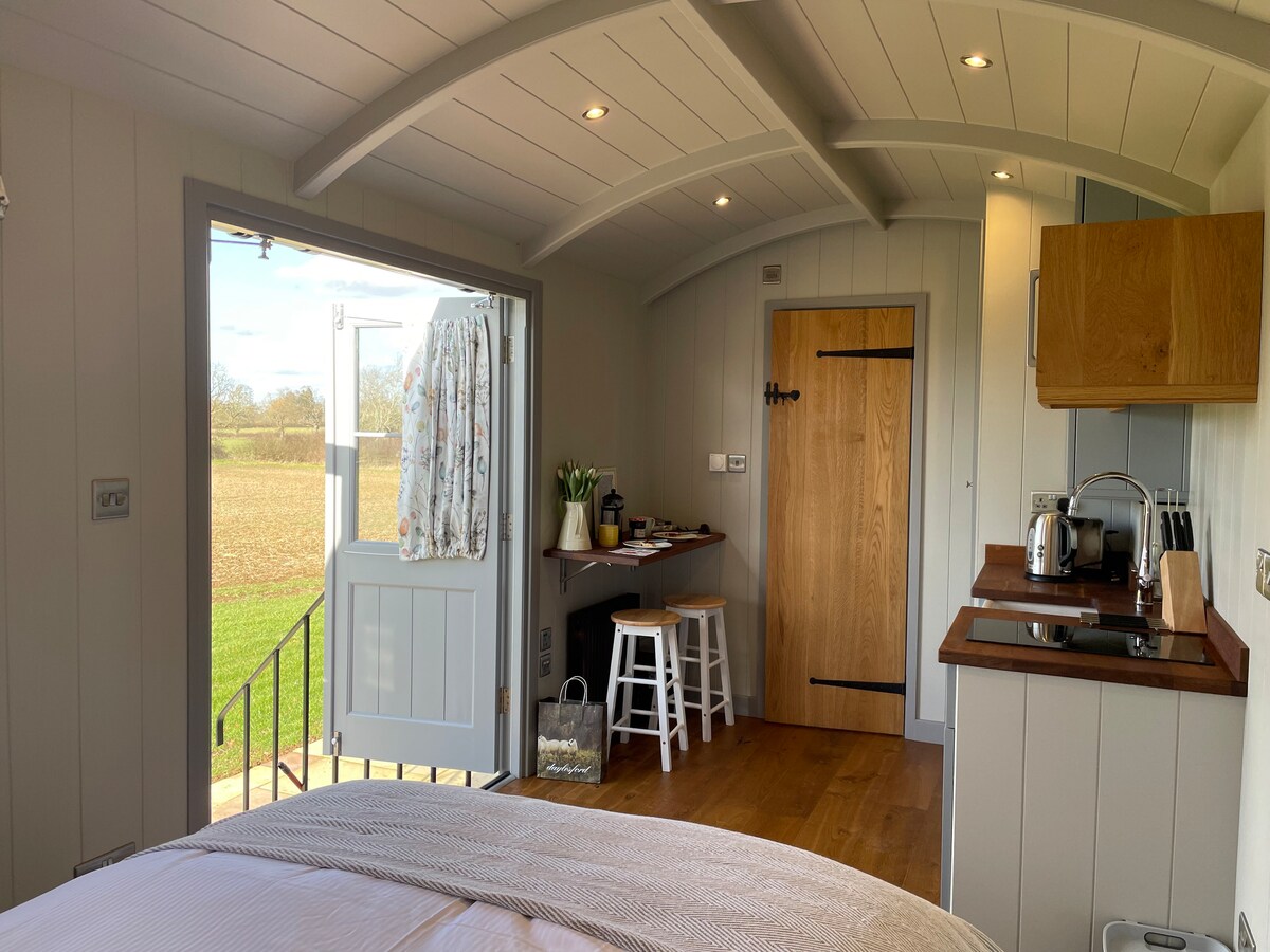 An open-plan living area is visible, featuring a spacious bed and a kitchenette. Natural light enters through the open door, which leads to an outdoor view of the tranquil field. A small dining space with stools and a wooden table is also present.