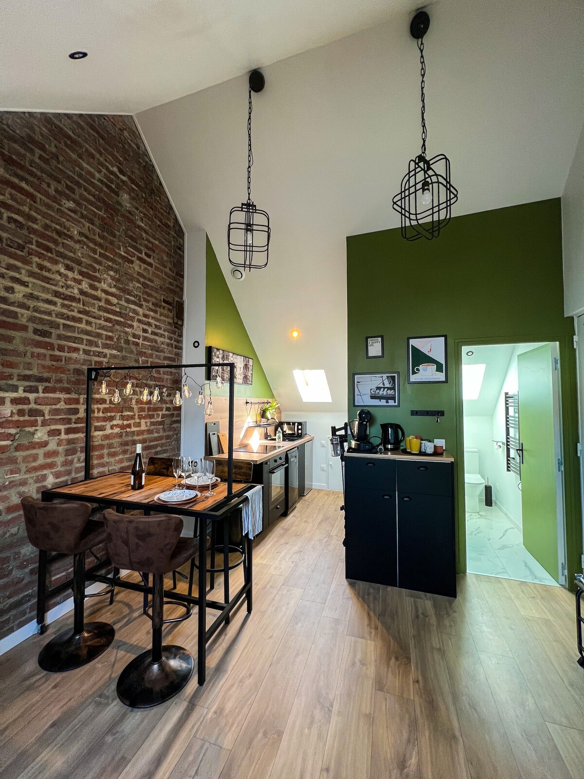 An open kitchen area features a mix of modern and rustic design elements, highlighted by exposed brick walls and green accent walls. A dining space with two bar stools is visible next to a kitchenette, illuminated by stylish pendant lighting.