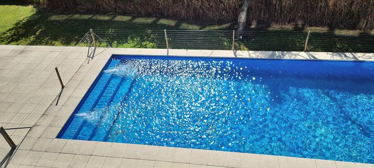 A long, modern swimming pool is visible, featuring crystal-clear water reflecting sunlight. Steps lead into the pool, surrounded by a spacious deck that is paved with light-colored tiles. Lush greenery can be seen in the background, enhancing the serene setting.