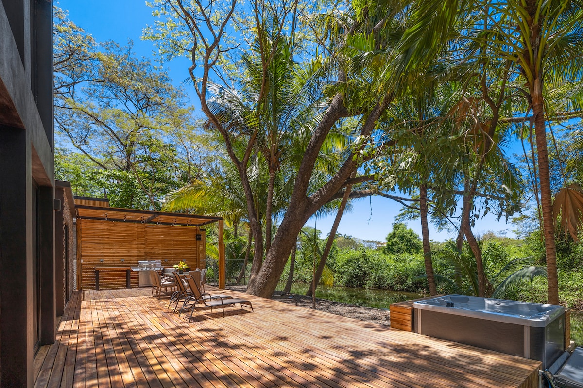 An expansive teak deck is surrounded by lush greenery and tall trees, providing a tranquil outdoor space. A hot tub is positioned near the edge of the deck, while lounge chairs are arranged for relaxation under the clear blue sky.