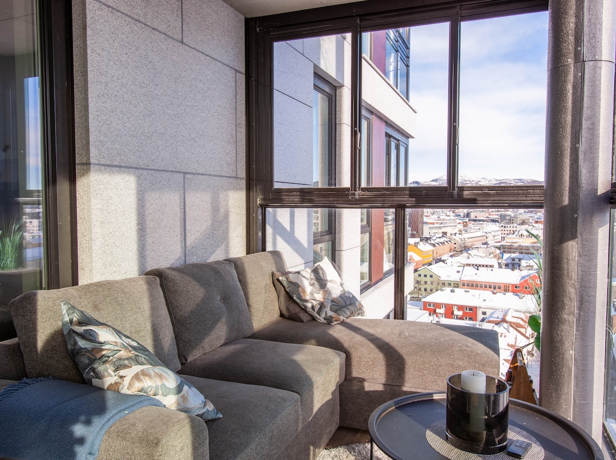 A modern seating area is positioned in a sunlit corner of the balcony, featuring a large, plush sofa adorned with decorative cushions. A small round table holds a candle, with expansive windows offering a view of the surrounding cityscape.