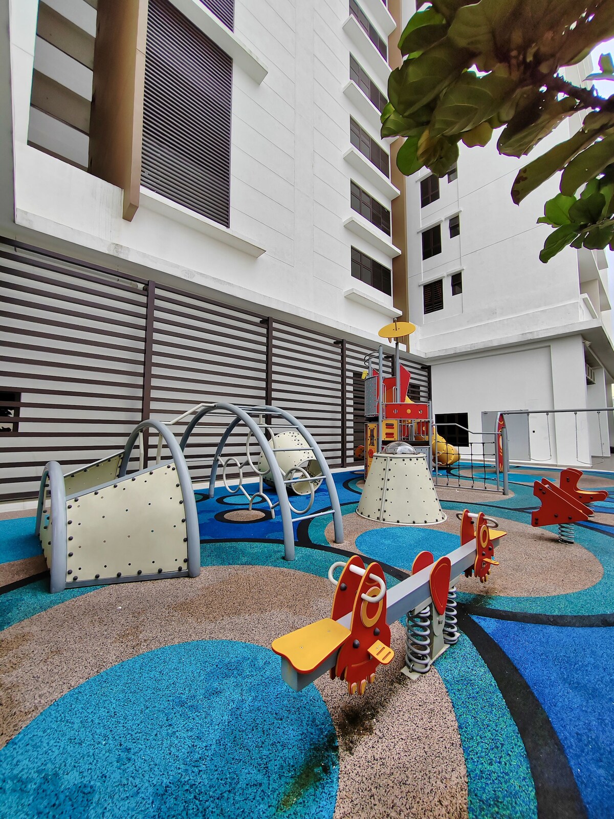 A colorful children's playground is displayed, featuring various play structures including slides, climbing frames, and swings. The ground is covered in blue and beige rubber flooring, offering a safe play area. Surrounding walls of the building are partially visible in the background.