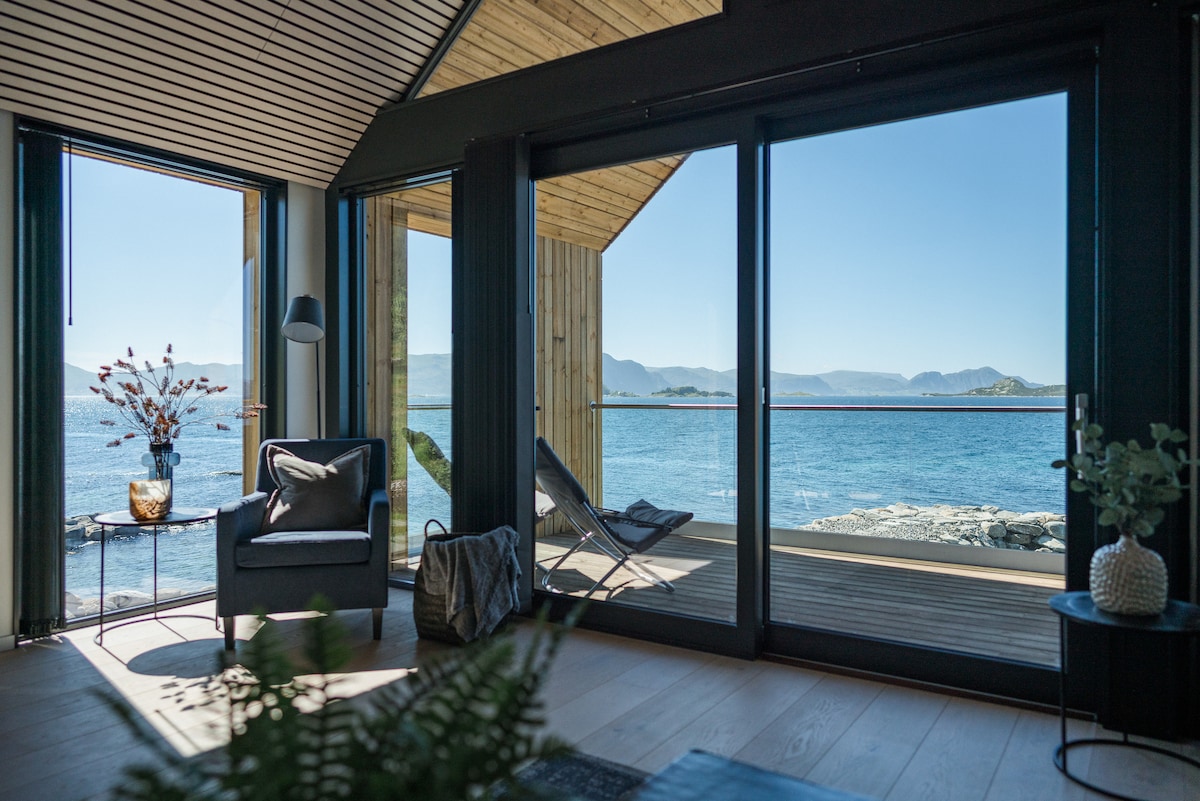 A modern living space features large glass doors that open to a terrace, showcasing panoramic views of the ocean and distant mountains. A comfortable chair sits beside a decorative plant and a side table, creating a serene spot for relaxation.