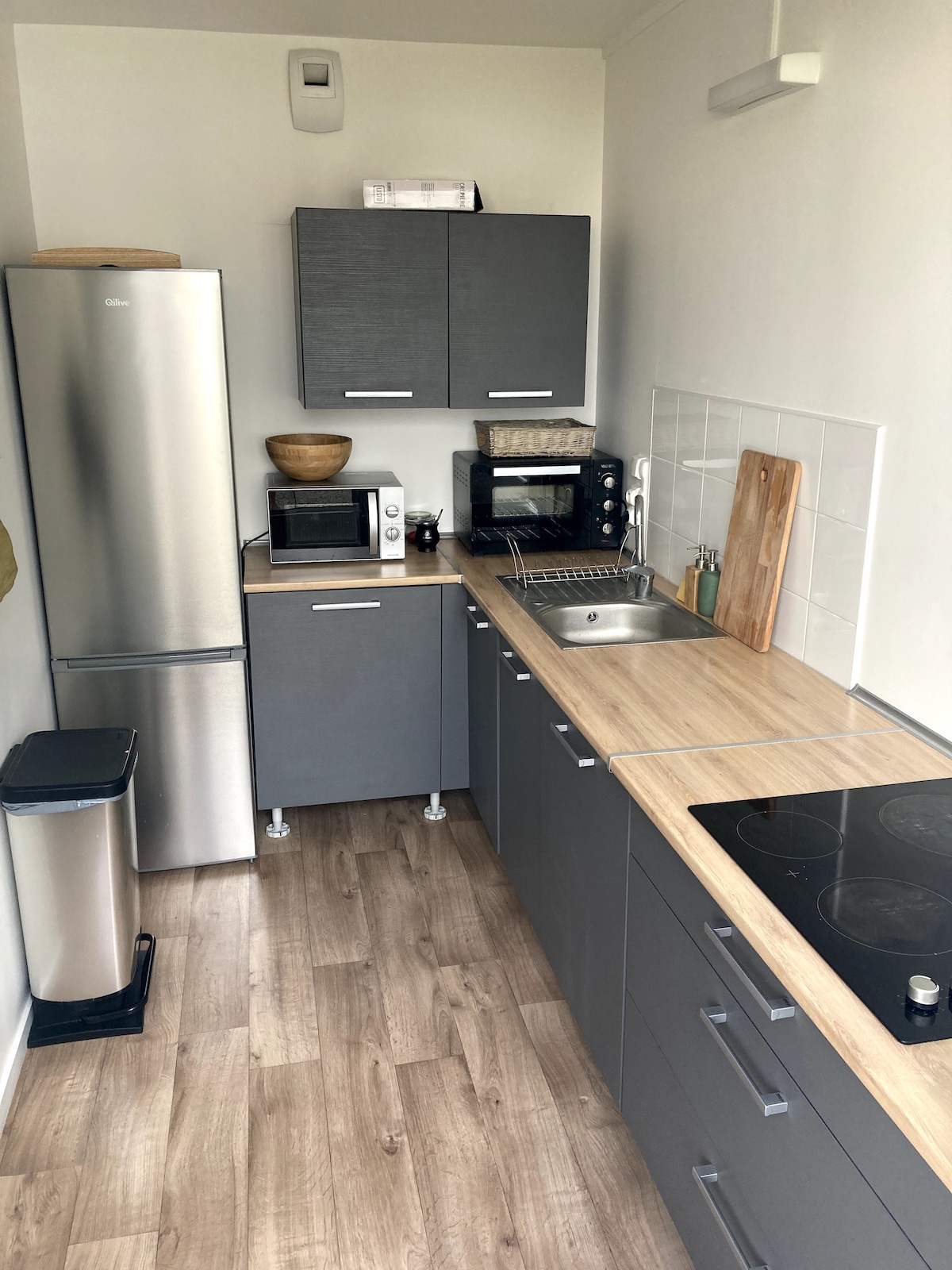 A modern kitchen is equipped with stainless steel appliances, including a refrigerator and microwave. Grey cabinetry complements the wood-tone countertops. Essential kitchen tools, such as a toaster and a kettle, are visible. Clear surfaces enhance the spacious feel of the area.