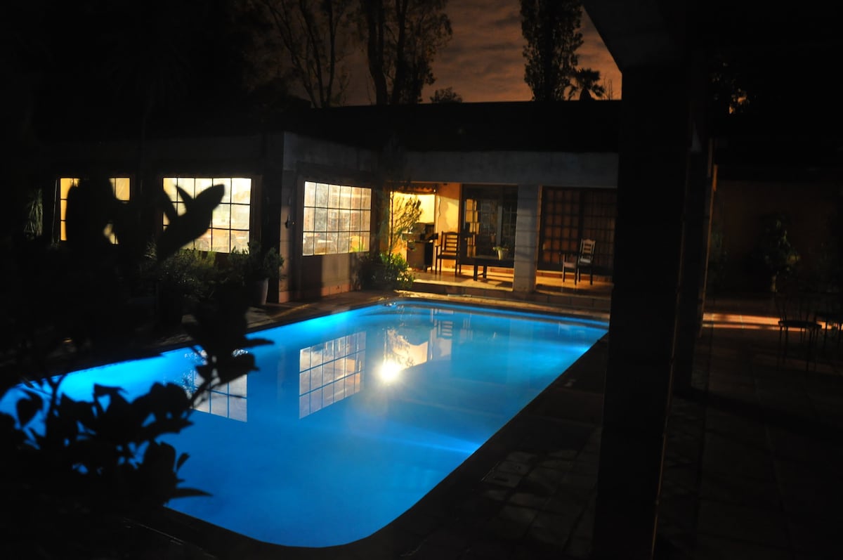 An illuminated pool reflects soft blue tones under the evening sky. Surrounding the pool, glass windows reveal a glimpse of the structure's interior, while outdoor seating arrangements are visible near the poolside, framed by lush greenery.