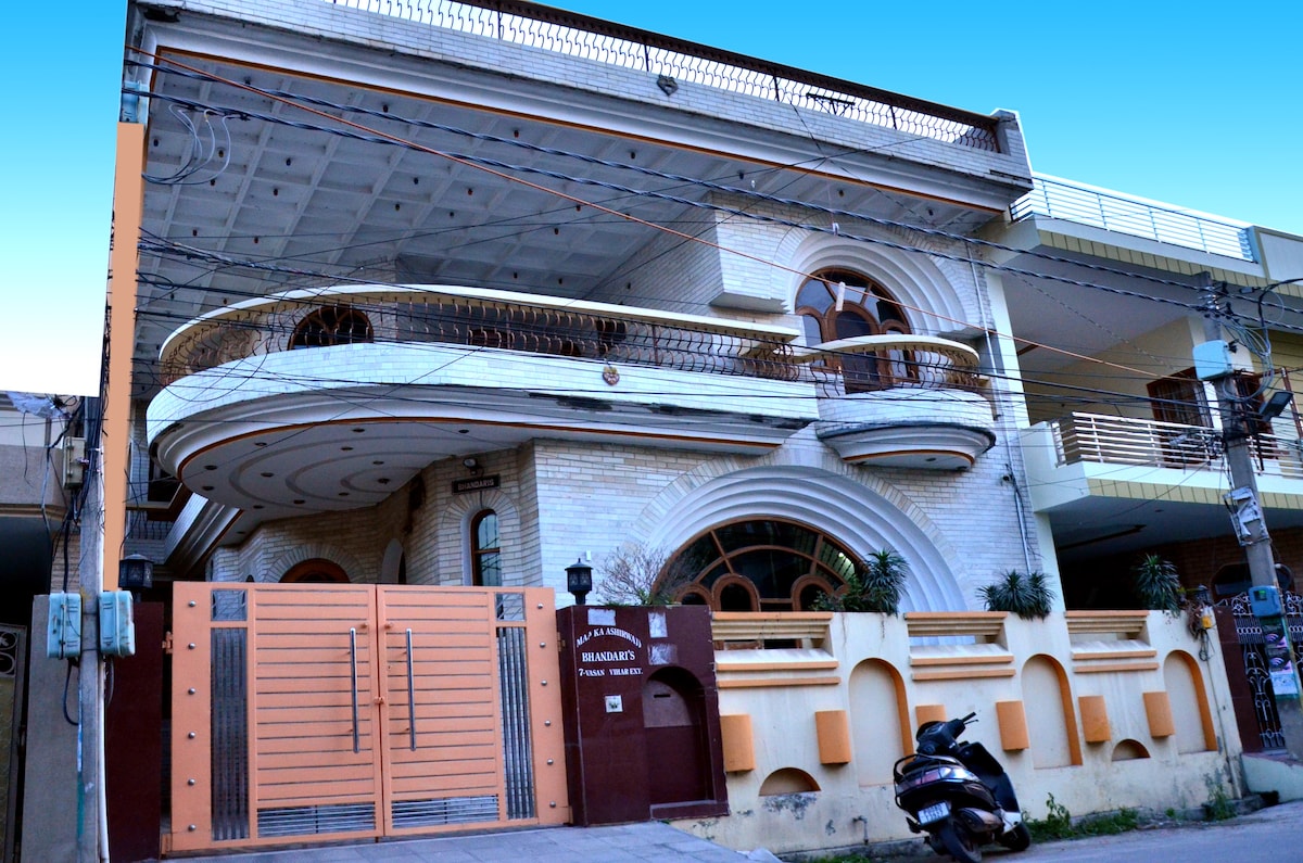 A three-story building features a decorative entrance with a large gate and an elegant facade. The architecture showcases curved balconies and arched windows, enhancing its spacious appearance. A motorcycle is parked in front, along with a well-maintained exterior and surrounding greenery.