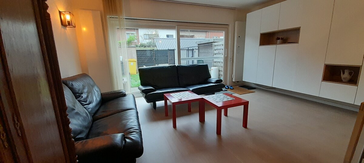 The living area features two black leather sofas facing a red coffee table, creating a comfortable space for relaxation. Large windows allow natural light to fill the room, while providing views of the garden. A minimalist white cabinet is positioned against the wall.