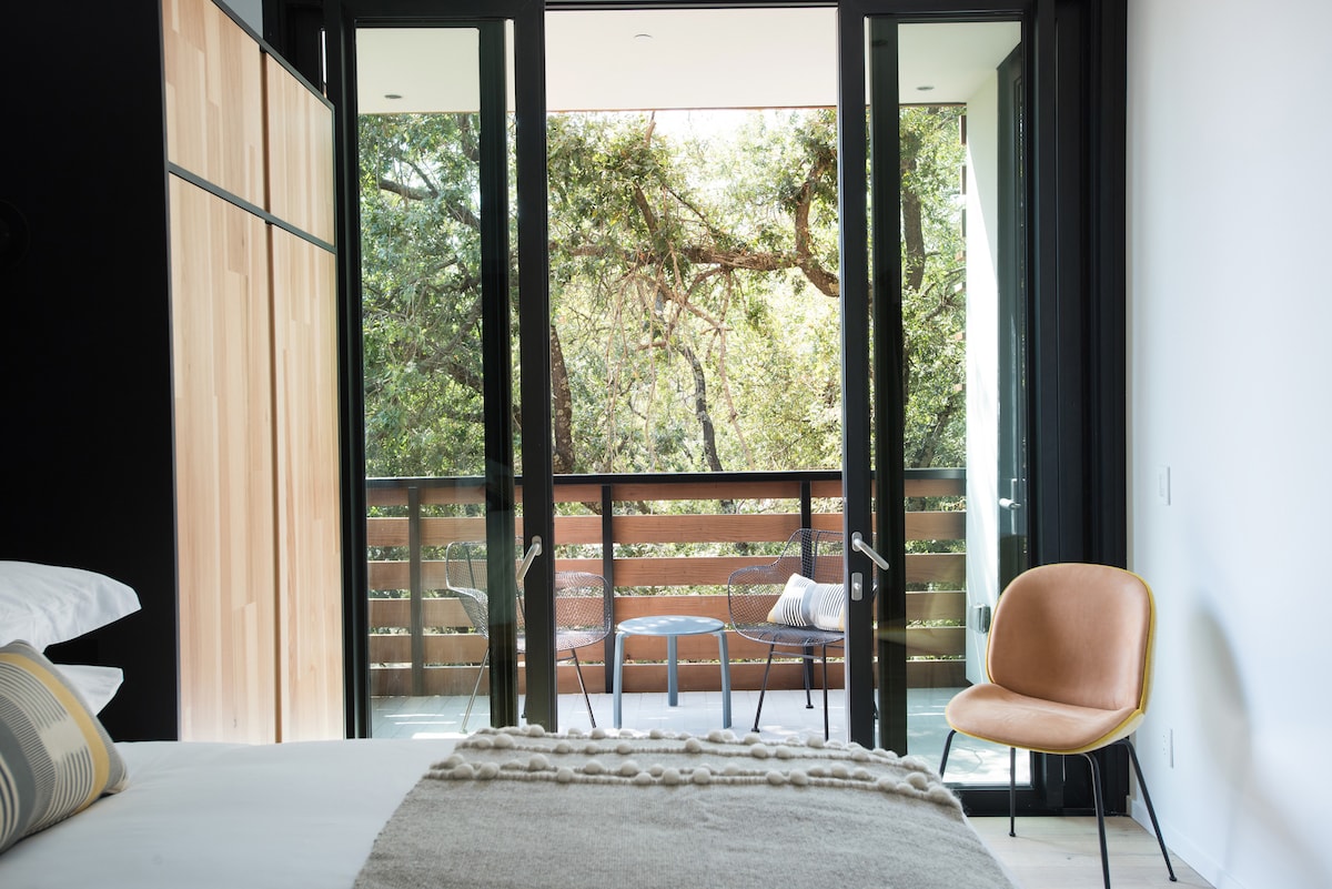 A spacious bedroom features large glass doors that open to a private balcony surrounded by greenery. A minimalist cream chair is positioned beside a small outdoor seating area, while soft linens and a decorative throw blanket adorn the inviting bed.