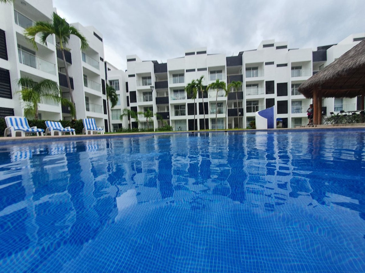 A clear view of a blue swimming pool reflecting white and black modern buildings. Loungers with blue and white stripes line the pool's edge, while palm trees add a touch of greenery to the serene outdoor space.