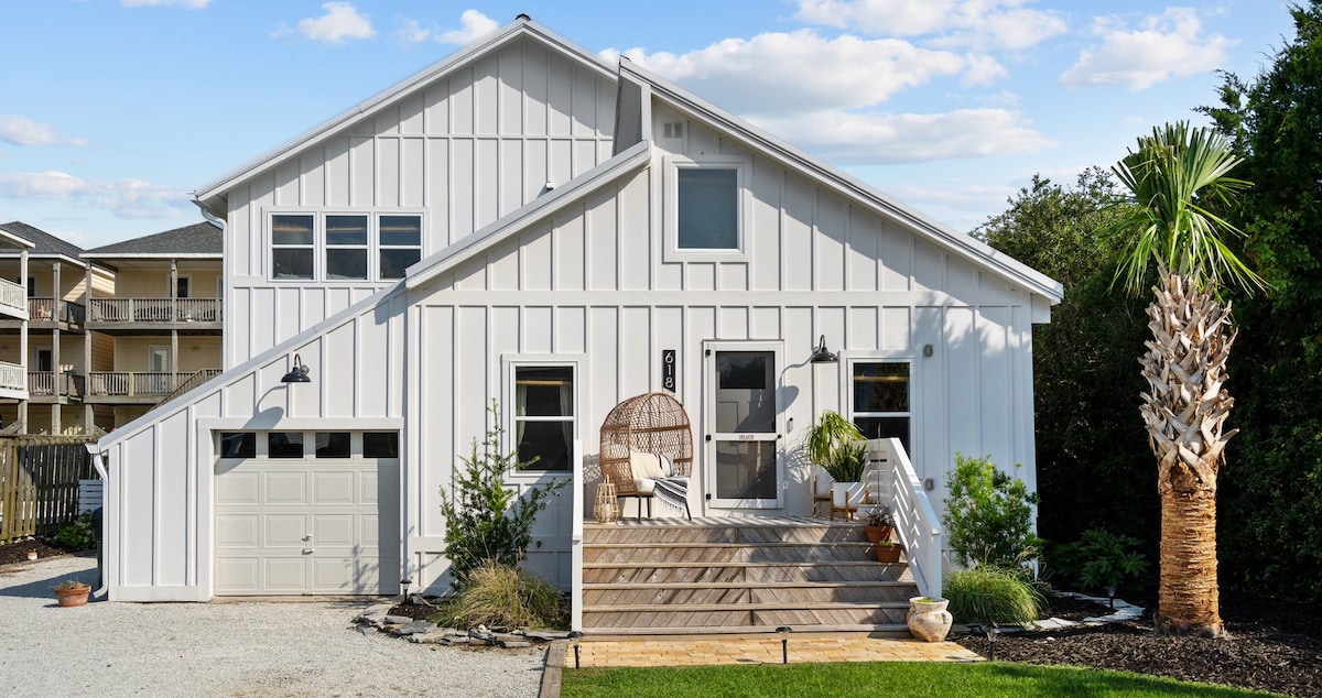 A modern exterior of a white bungalow is shown, featuring a landscaped entrance with wooden steps and potted plants. A circular lounge chair is positioned to the side, and tall palm trees frame the property while a gravel driveway leads to the garage.