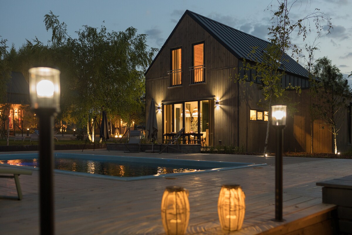 A modern wooden structure is illuminated at dusk, showcasing large glass doors that invite guests into the interior. Soft lights are placed around a wooden deck, and a clear swimming pool reflects the evening sky. Lanterns add a warm glow to the outdoor space.