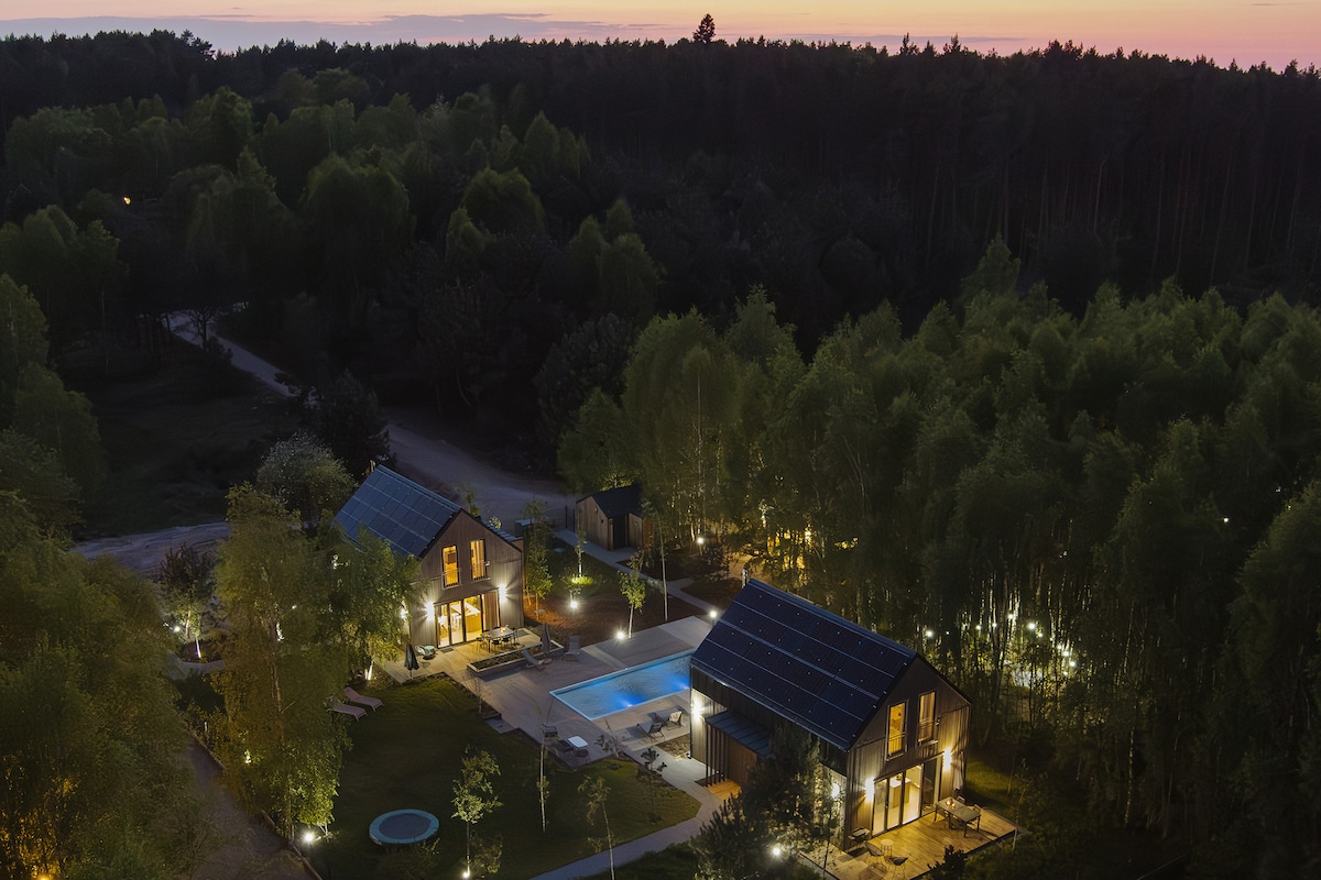 An aerial view of the eco-friendly homes reveals a serene landscape surrounded by tall trees. The outdoor area includes a well-lit swimming pool, lounge chairs, and a spacious garden, creating a peaceful retreat at dusk.