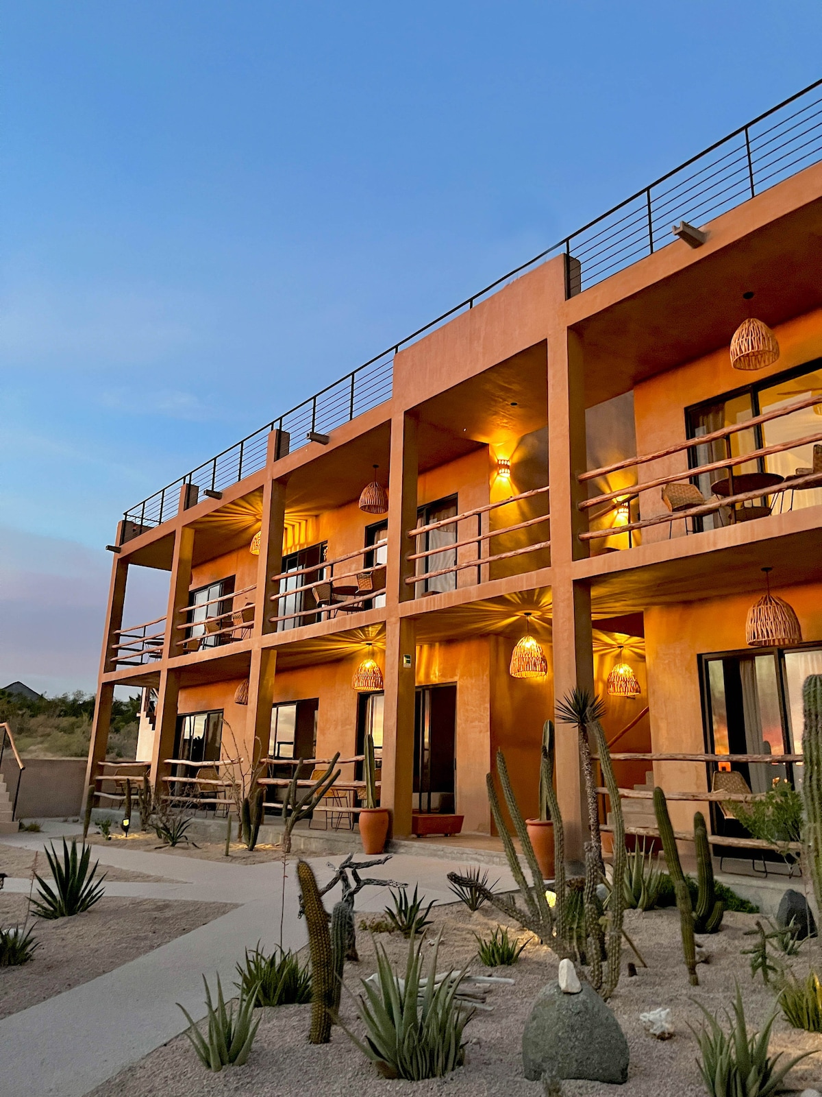 The exterior of a boutique hotel is showcased, featuring a two-story structure with warm lighting illuminating the balconies. Cacti and desert flora surround the pathways, enhancing the serene desert landscape as the evening sky transitions to twilight.