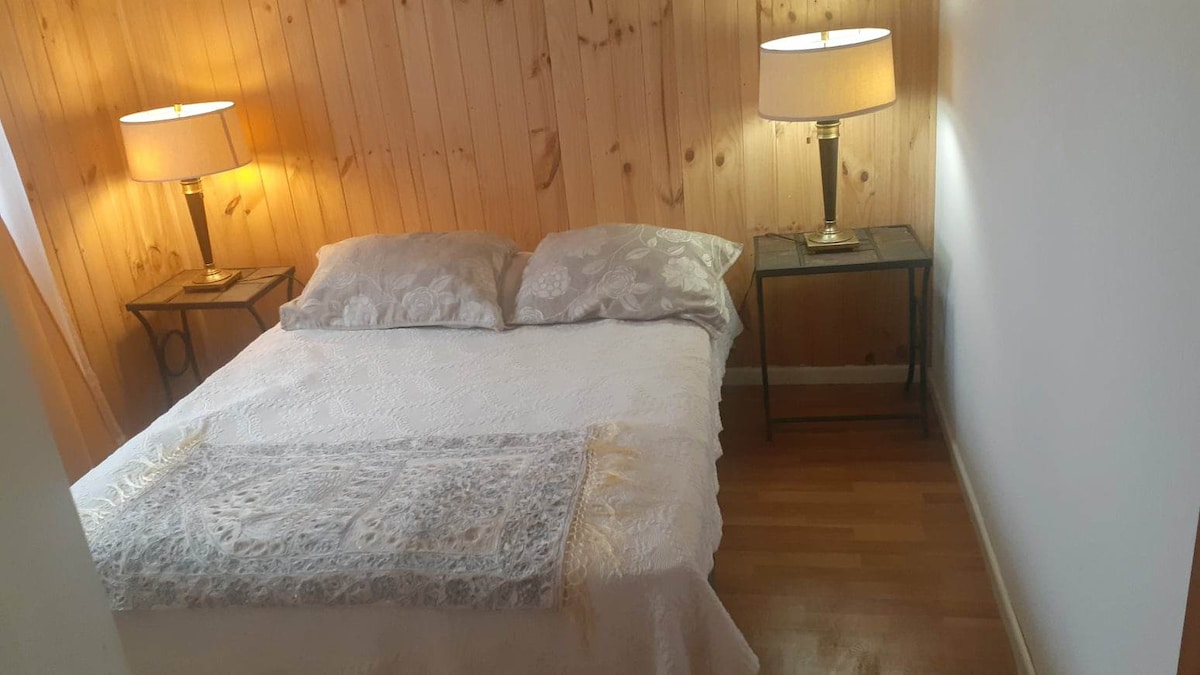 A cozy sleeping area features a spacious bed dressed in light bedding. Matching bedside tables are placed on either side, each holding a lamp that softly illuminates the wooden walls, contributing to a serene and relaxing environment.