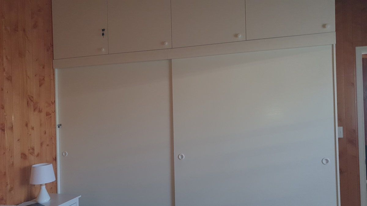 A sliding wardrobe with two large doors, accompanied by minimalistic upper cabinets in a light color, is situated against a wooden wall. A small bedside lamp is visible in the foreground, providing illumination.