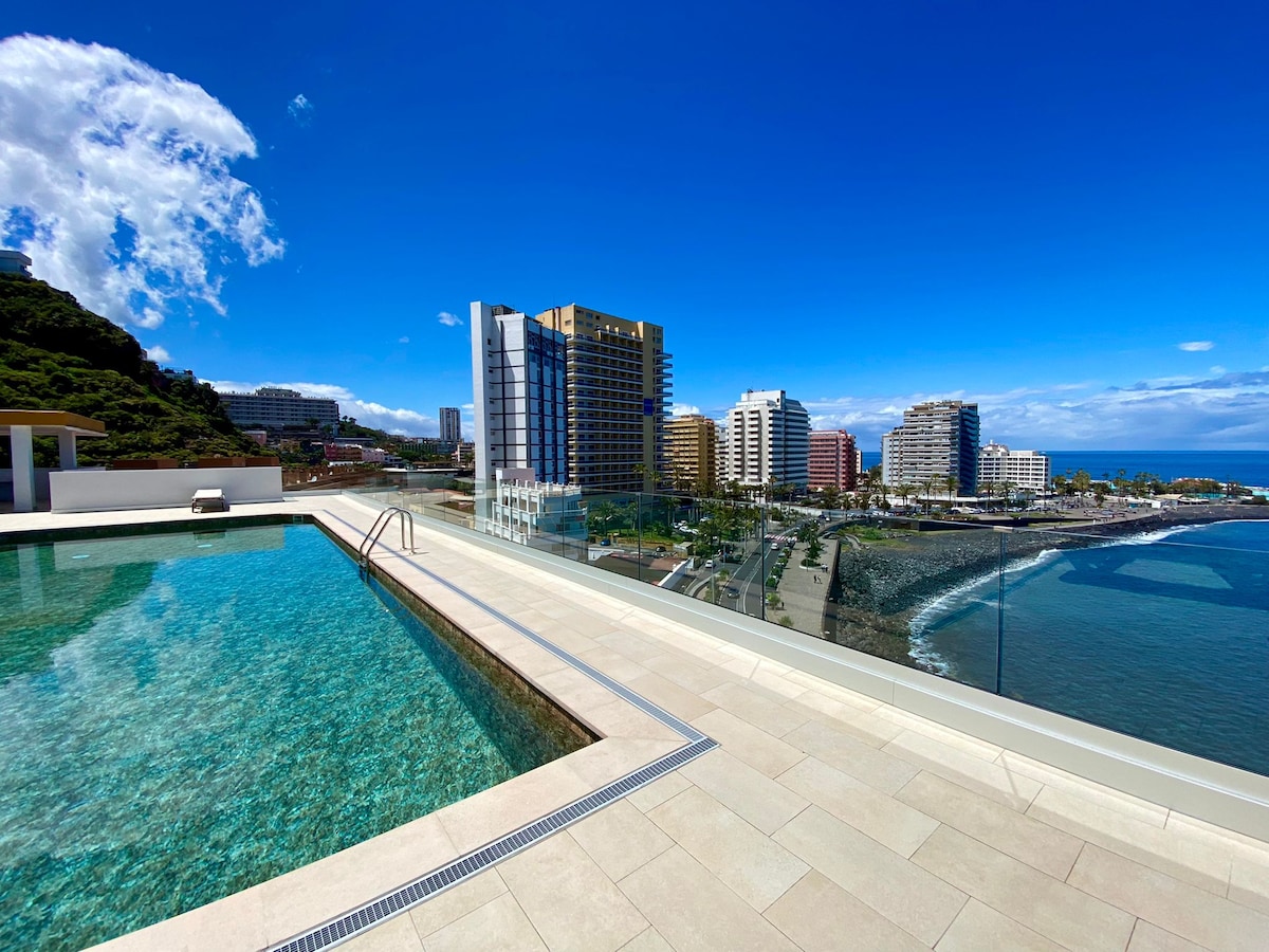 A rooftop pool is viewed against a backdrop of the Atlantic Ocean and surrounding buildings. The clear water of the pool reflects the blue sky and clouds, while a paved area with lounge chairs extends beside it. The serene atmosphere invites relaxation.