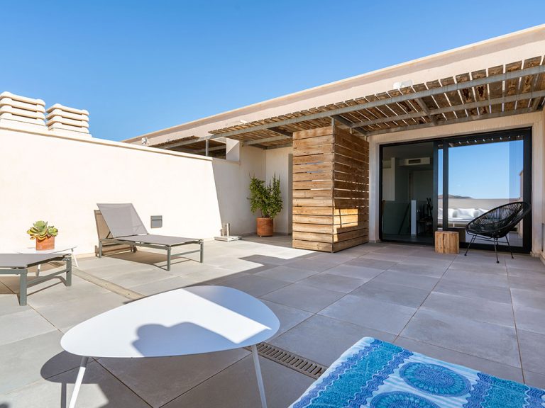 A private terrace features modern outdoor furniture, including lounge chairs and a small table. Smooth tiles line the area, with a wooden partition adding privacy. Potted plants provide a touch of greenery, and large glass doors lead into the interior of the duplex.