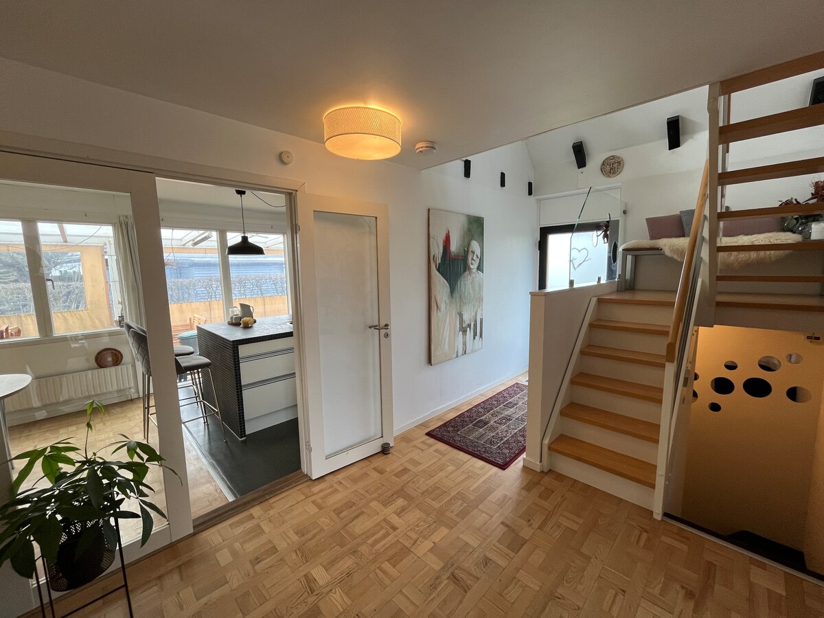 An entryway features a modern light fixture and a colorful artwork on the wall adjacent to the staircase. The wooden stairs lead to a cozy sleeping area, while an open space reveals views of the kitchen and outdoor landscape through large glass doors.