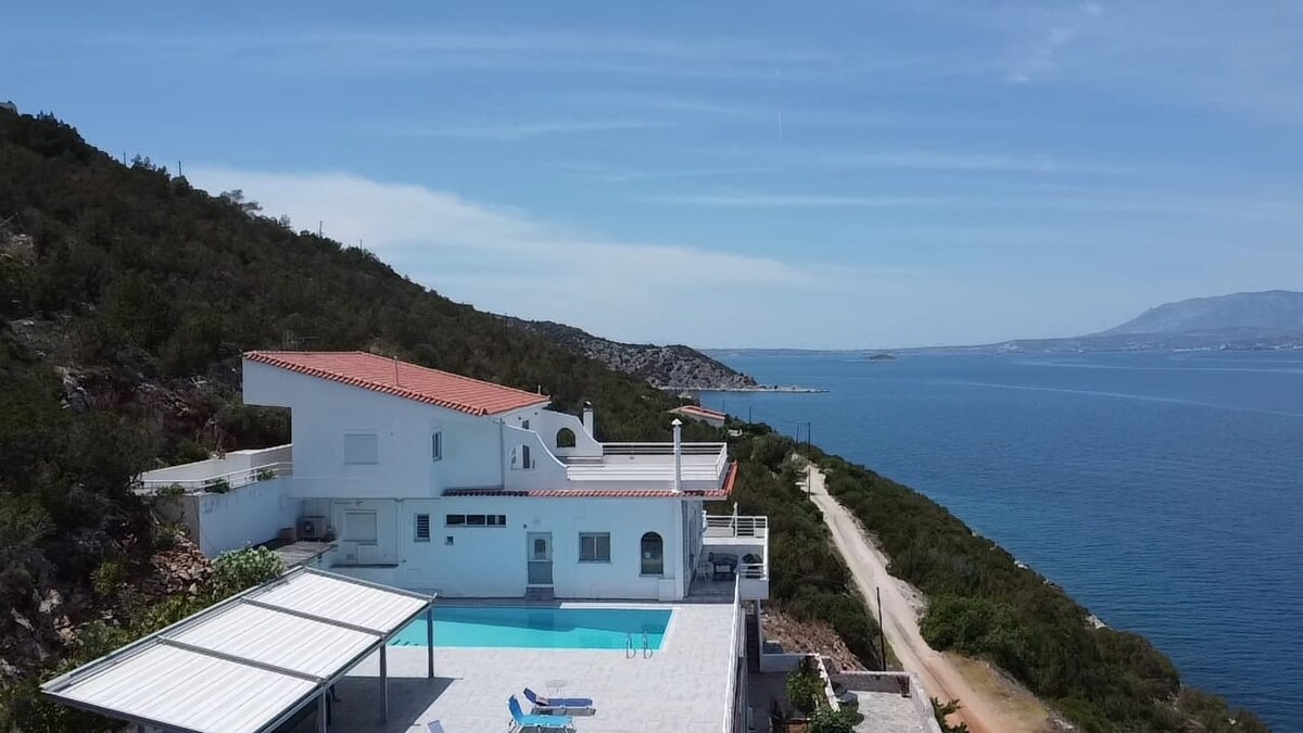 The villa is presented from an aerial perspective, showcasing its modern architecture and spacious exterior. A sea water pool is visible, reflecting the surrounding blue waters. The property is nestled against a backdrop of lush greenery and the coastline, emphasizing its serene setting.