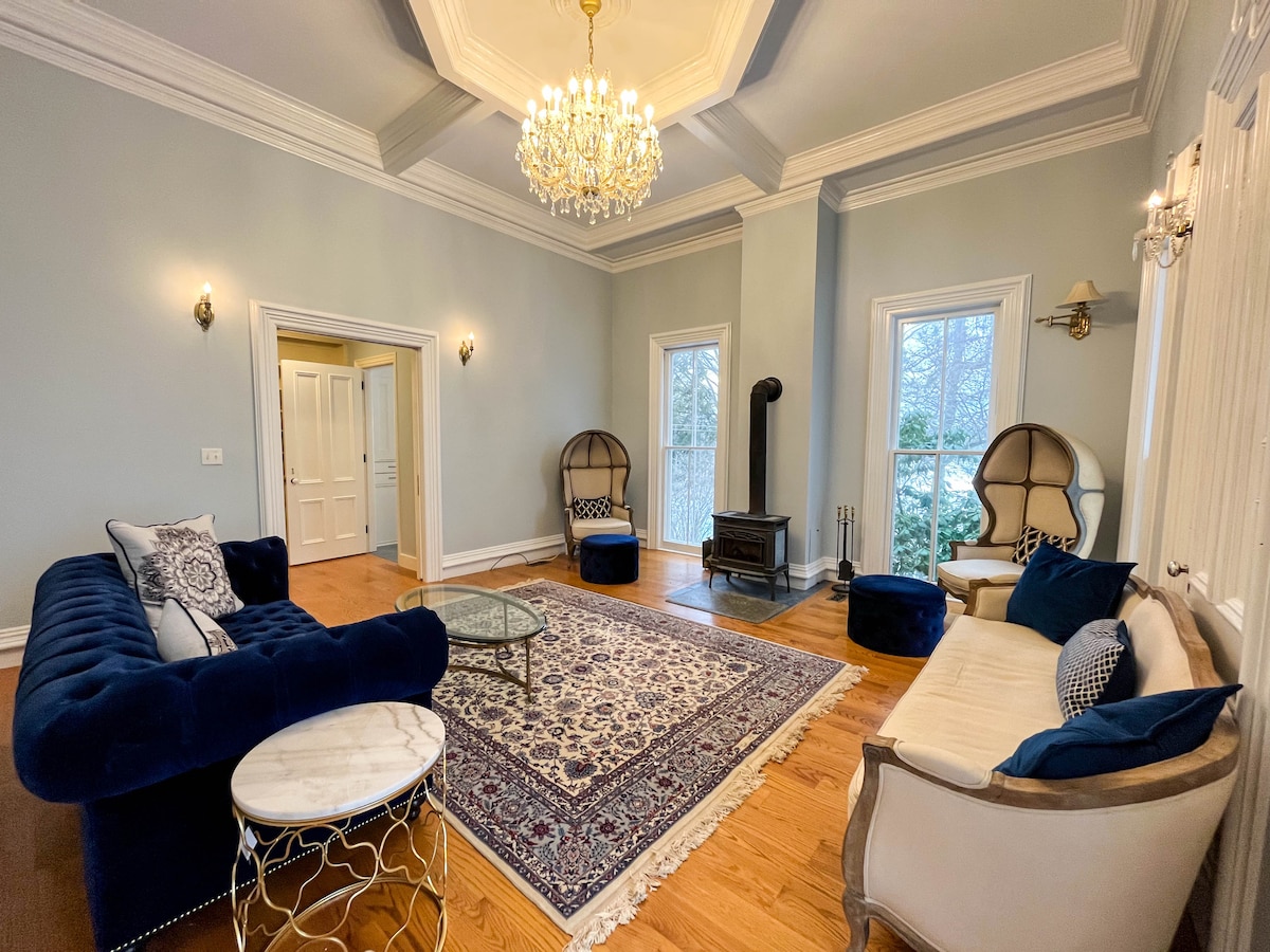 A spacious living room features a crystal chandelier hanging from the coffered ceiling and large windows offering views of the exterior. Elegant seating arrangements include a plush blue sofa, two armchairs, and a light-colored settee, complemented by a decorative rug and a wood-burning stove.