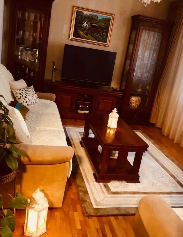 A comfortable living area is highlighted by a plush sofa, accompanied by a wooden coffee table. A television is positioned on a cabinet, while two glass display cases feature decorative items. Soft lighting from lanterns and natural light from the window create a warm ambiance.