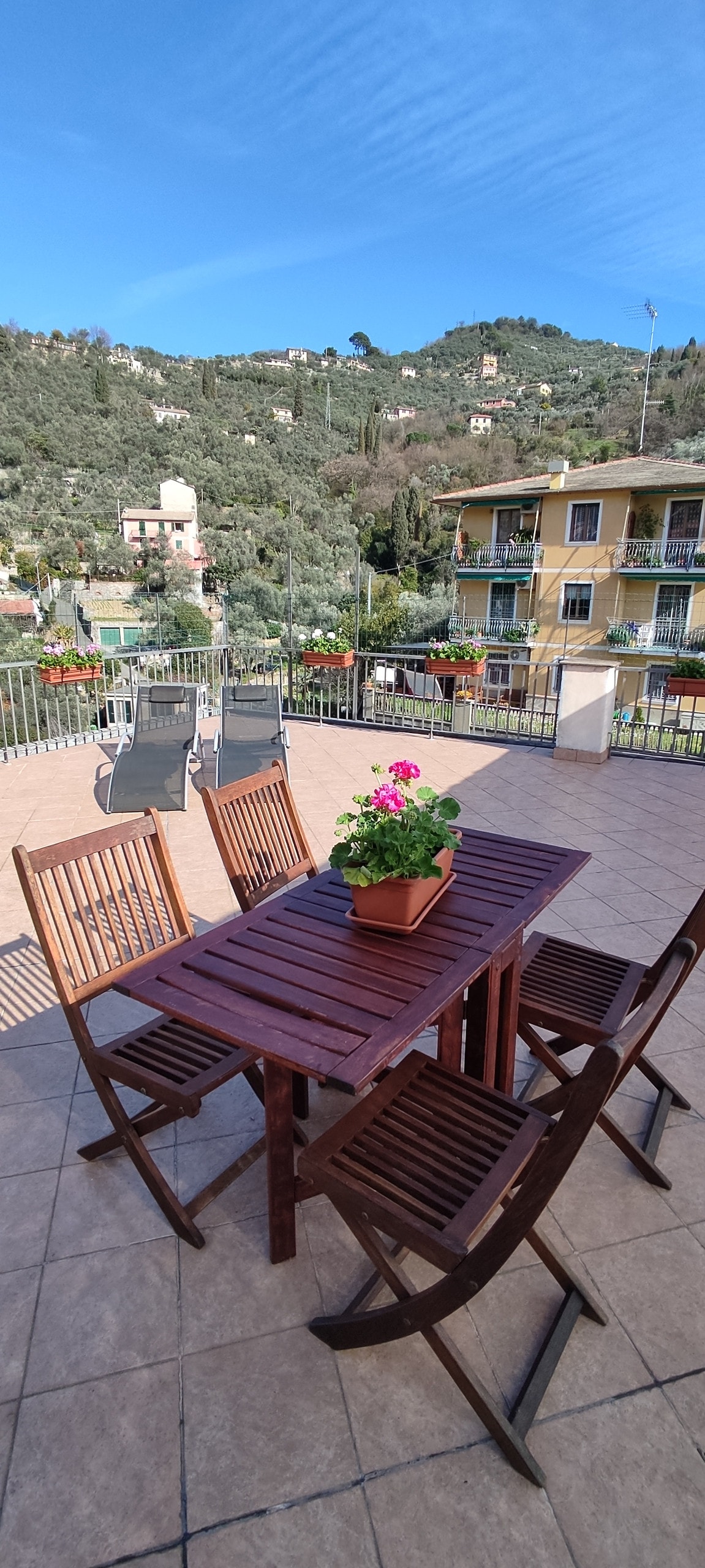 A spacious terrace features a wooden dining table surrounded by four folding chairs. A potted plant adds a touch of color, while scenic hills and greenery are visible in the background. This outdoor area is designed for relaxation and dining al fresco.