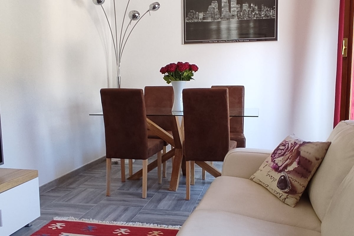 A dining area features a glass table surrounded by four brown chairs, creating a modern yet inviting space. A vase with roses adds a touch of color. A plush sofa is positioned nearby, providing comfort and accessibility in the living area.