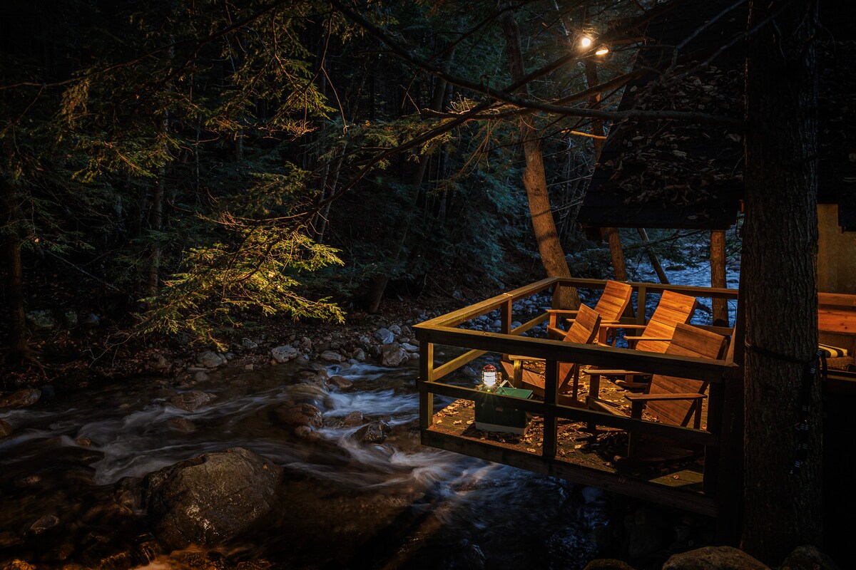 A secluded screened cabin is positioned over a gently flowing brook, surrounded by dense trees. Wooden chairs are placed on the deck, inviting relaxation in nature. Soft lighting from above contributes to the serene ambiance of the evening scene.