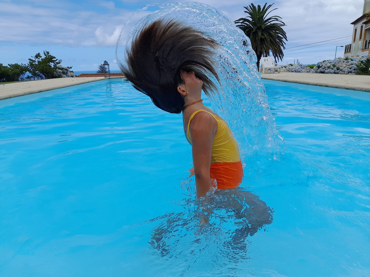 A person is seen in mid-motion, creating splashes in a clear blue swimming pool. The pool reflects the sky above, and palm trees can be seen in the background, contributing to a relaxed outdoor setting.