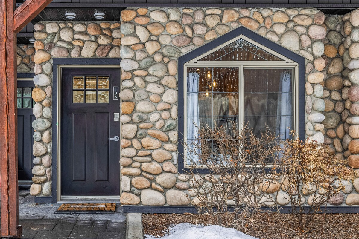 A charming stone exterior features a dark-colored door framed by an arched window. The window showcases warm indoor lighting, while snow gently blankets the ground surrounding decorative shrubs. The natural stone highlights the rustic character of the entrance.