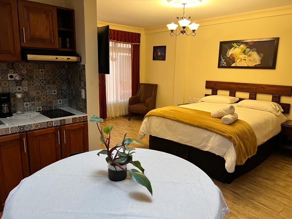 A well-equipped living space is presented with a spacious bed covered in a soft yellow blanket. A small round table with a potted plant is visible in the foreground. A cozy armchair and a wall-mounted television are situated near a window with light drapes, enhancing the room's functionality.