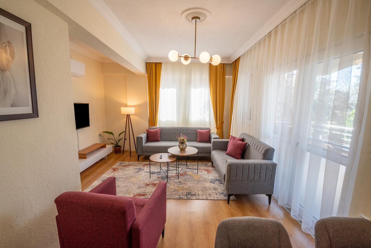 A bright living area features large windows draped with sheer curtains, allowing natural light to fill the space. Two sofas and two armchairs are arranged around a circular table, complemented by decorative pillows. A wall-mounted TV is situated above a minimalist shelf on one side.