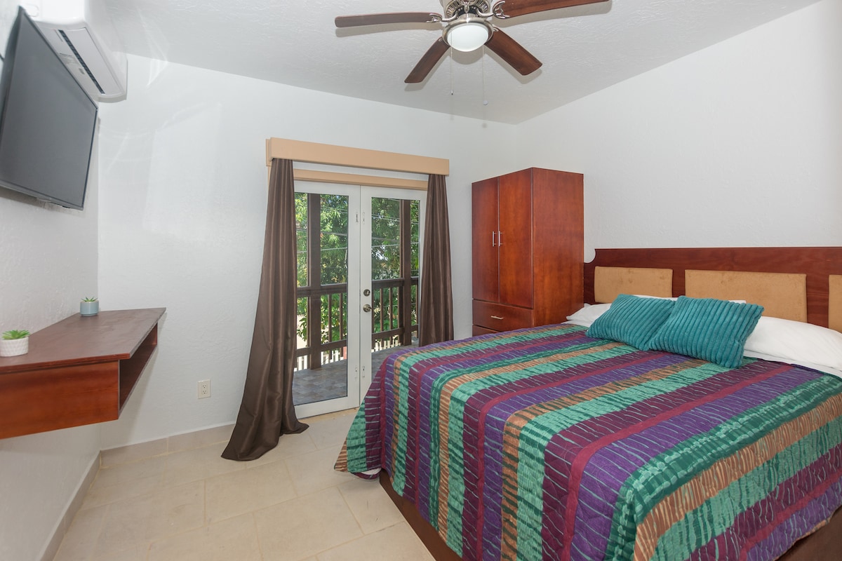 The guest bedroom features a spacious queen bed adorned with a colorful striped blanket. A flat-screen Smart TV is mounted on the wall, while a ceiling fan provides comfort. Sliding glass doors lead to a private balcony, enhancing the room's bright atmosphere.