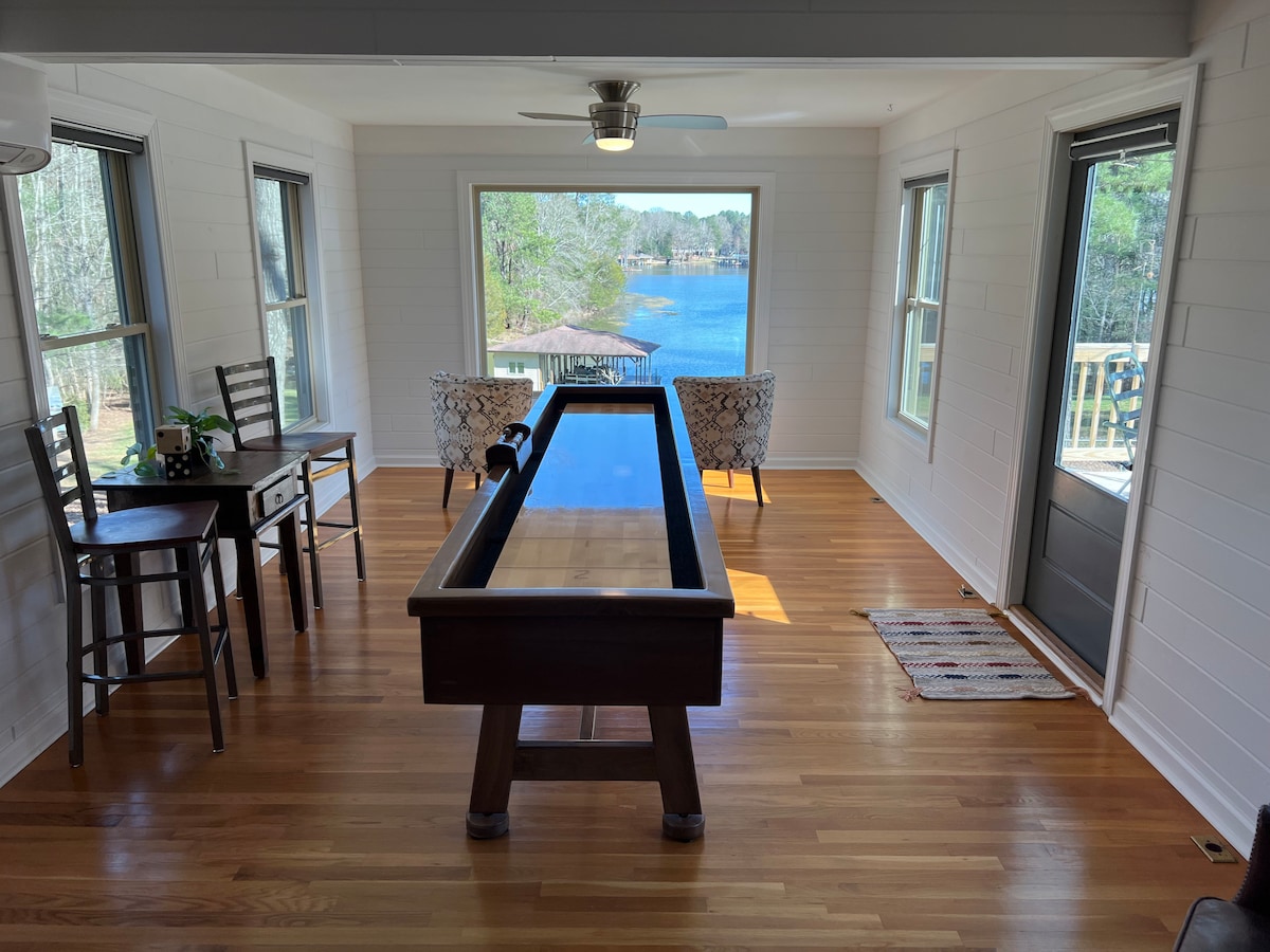 A game room features a full-size shuffleboard table at its center, surrounded by two patterned chairs and a dining table with two stools. Large windows allow natural light to flood the space and offer scenic views of the lake and private dock.