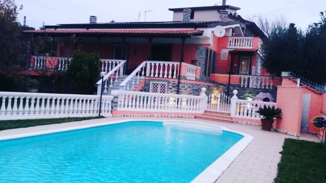 A private pool is framed by manicured grass and surrounded by a white balustrade. The two-story villa, with a terracotta roof, is visible in the background, showcasing multiple sets of stairs leading up to its entrance.