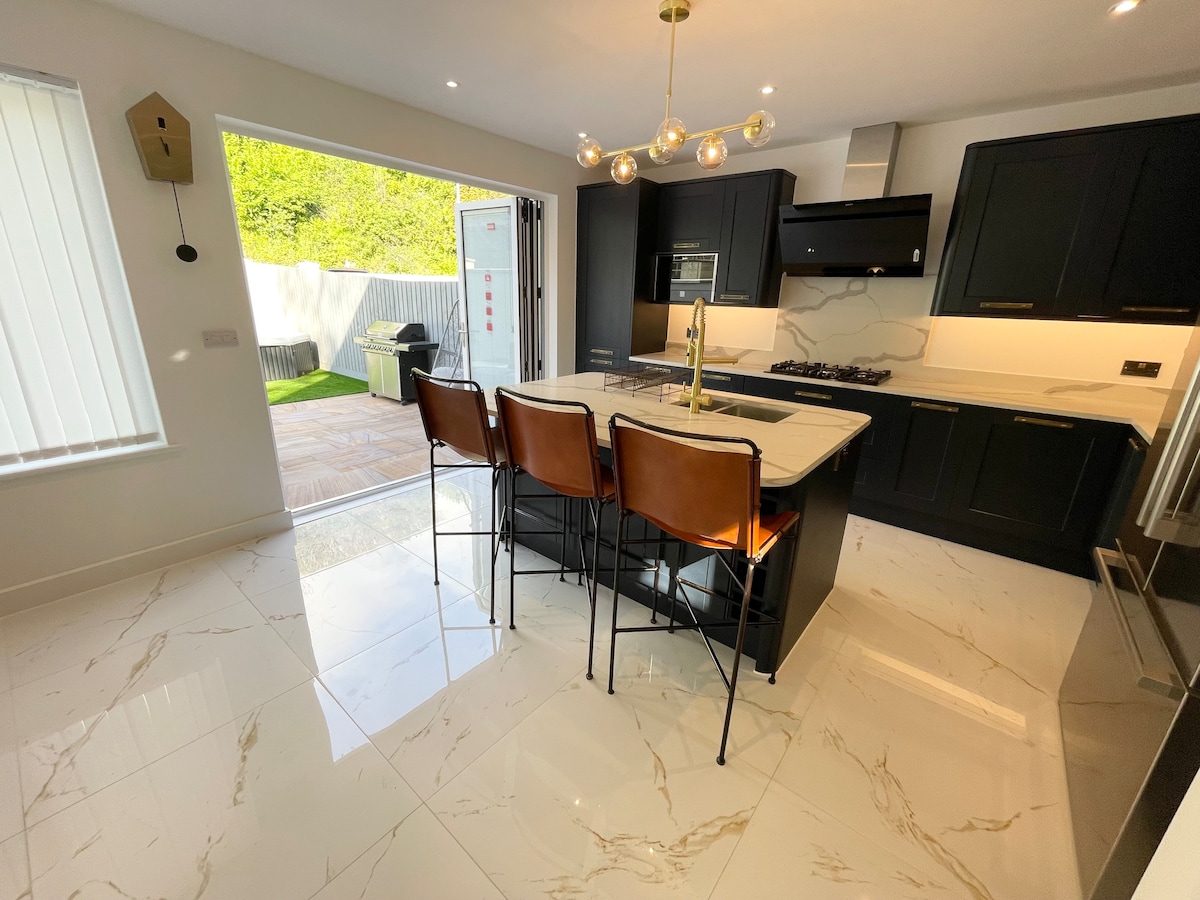 The contemporary kitchen features sleek black cabinetry and a spacious island with seating for four. Bi-fold doors open to the garden, allowing natural light to fill the room. Modern appliances, including a double oven and a polished countertop, enhance the cooking space.