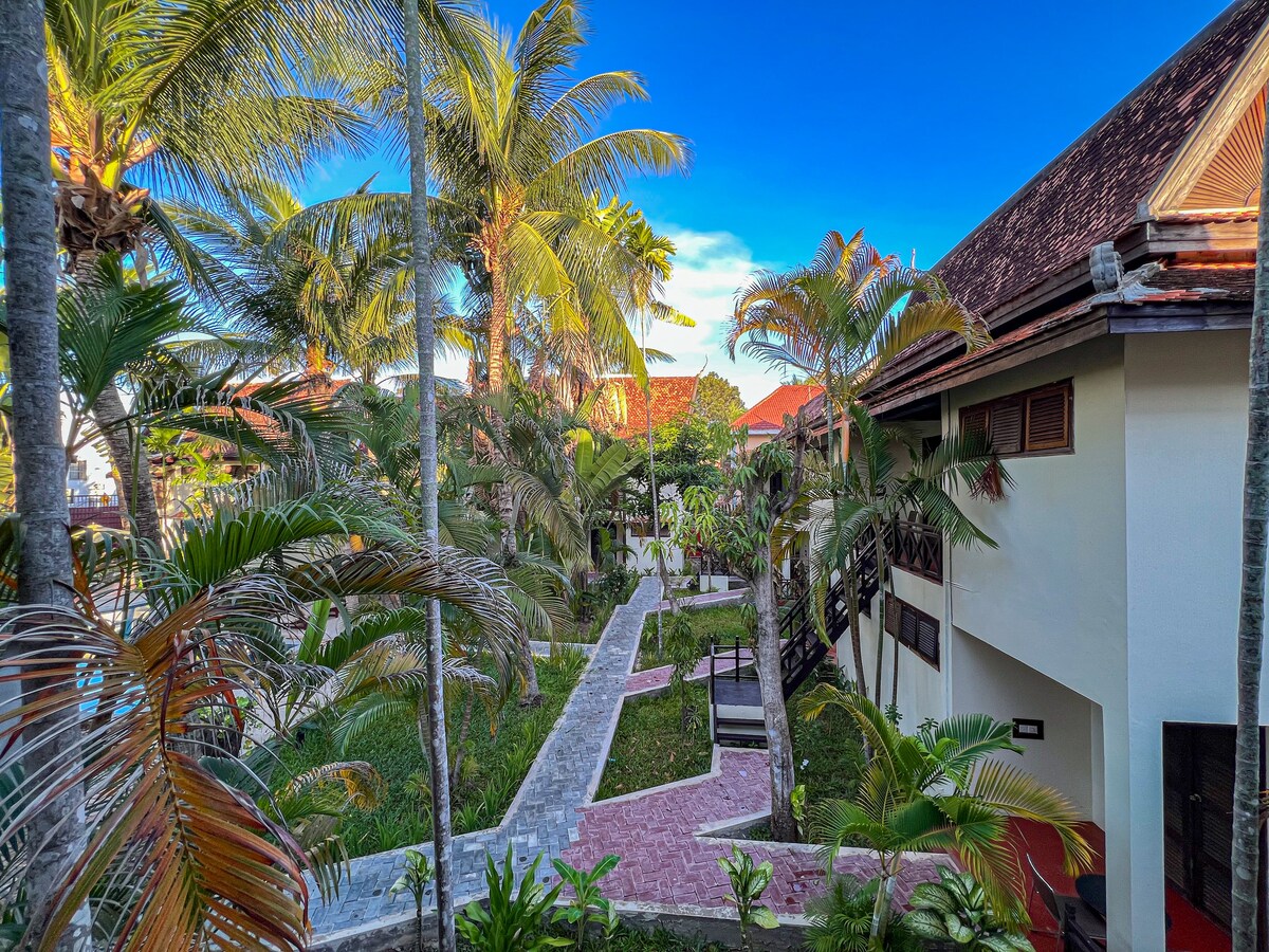 Lush tropical landscaping surrounds the pathway that winds through the property. Palm trees frame the view of the buildings, while vibrant greenery is visible on either side. A blue sky with wispy clouds completes the serene outdoor setting.