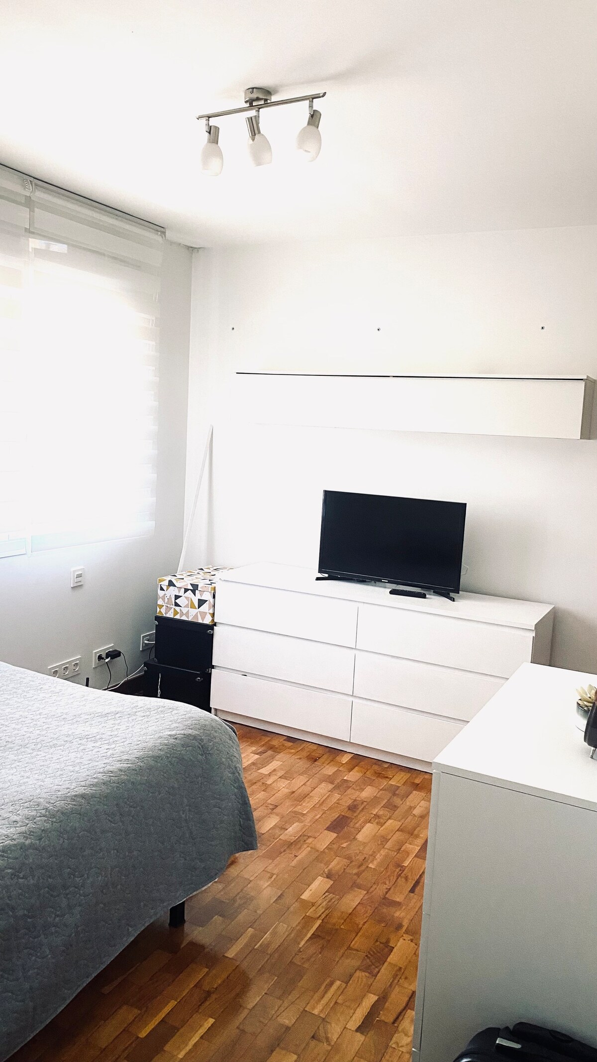 A bright bedroom features a spacious bed draped with a light gray blanket. A sleek white dresser and a small television are positioned against the wall. Natural light filters through the window with light-filtering shades, creating a calm and inviting environment.