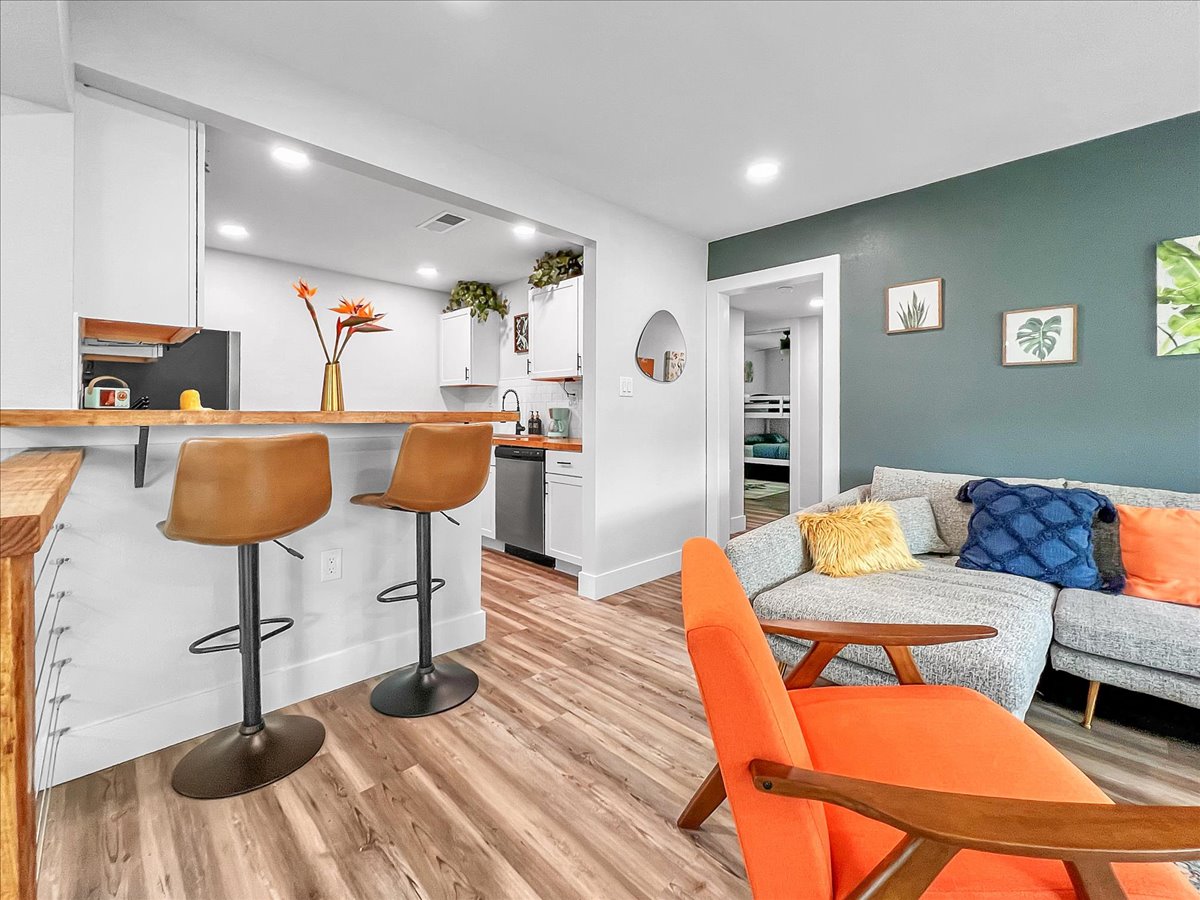 A modern living area features a cozy sectional sofa with colorful cushions and a vibrant orange chair. A breakfast bar with two stools is visible, accompanied by stylish kitchen amenities in the background. Soft lighting enhances the inviting space, with tasteful wall decor adding character.