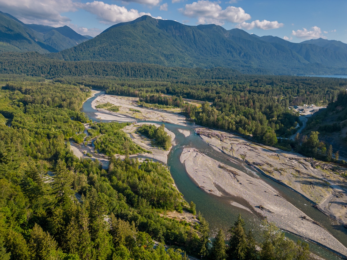 Aerial view of a river winding through a lush landscape, framed by rolling mountains in the distance. Sandy banks contrast with the vibrant green foliage along the riverbanks. The scene captures the serene beauty of nature, showcasing textures and patterns created by the land and water.