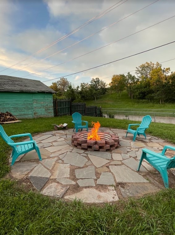 A circular flagstone patio surrounds a vibrant fire pit, with flames flickering at the center. Four aqua-colored chairs are placed evenly around the fire pit, inviting relaxation. The backyard features lush grass and a scenic backdrop of greenery and trees.