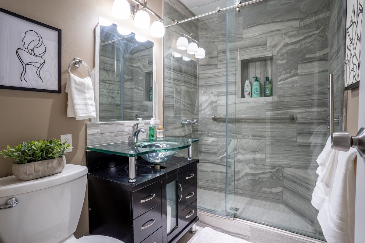 A modern bathroom features a walk-in shower with a glass enclosure, showcasing textured gray tiles. A sleek black vanity with a glass sink is complemented by a wall-mounted mirror, providing ample space for toiletries. Soft lighting adds clarity to the organized space.