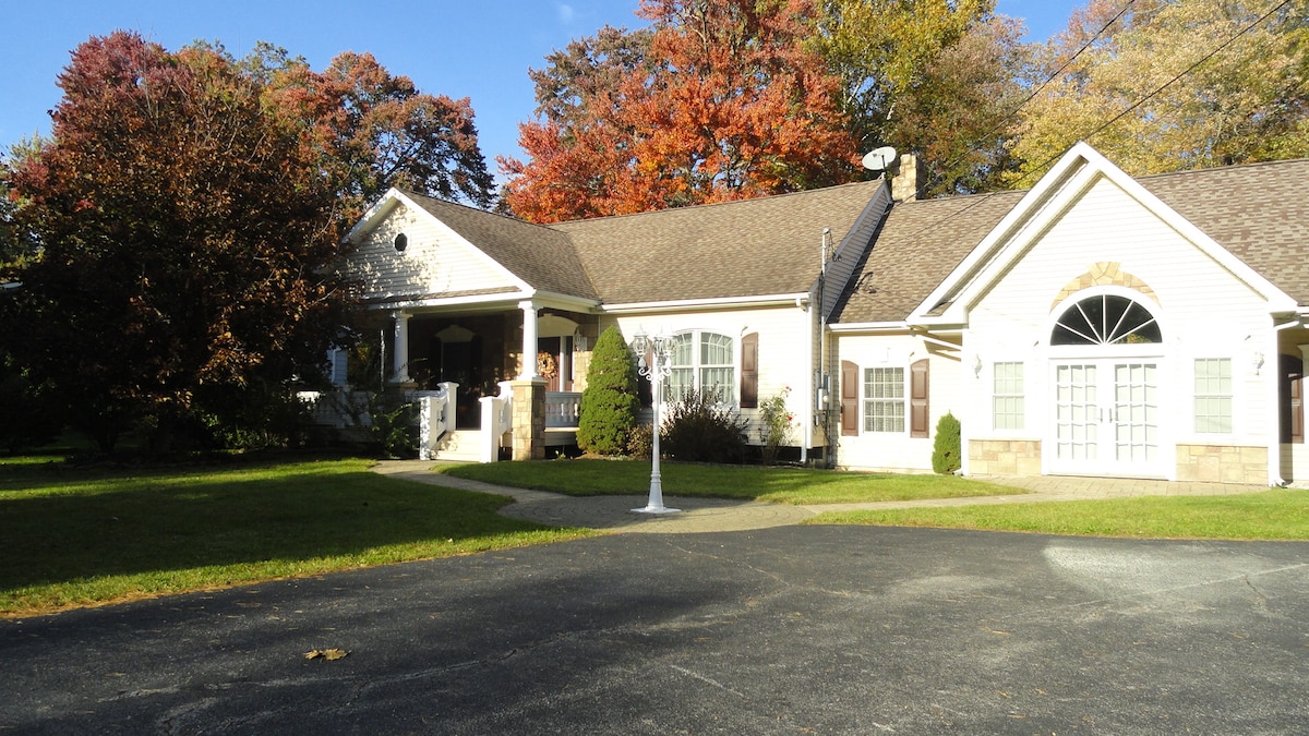 A spacious home sits surrounded by colorful trees in autumn. The house features a front porch with a welcoming entrance and large windows, allowing natural light to fill the interior. A basketball hoop stands in the driveway, accommodating outdoor activities.