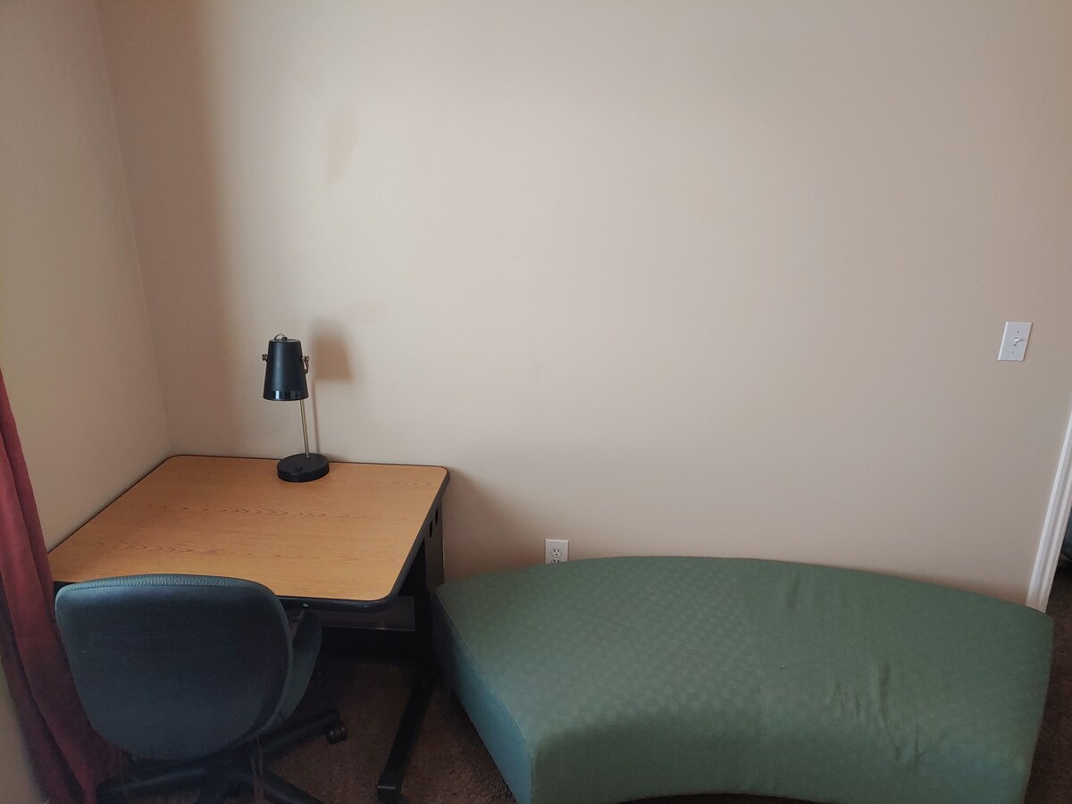 A small workspace is featured with a wooden desk and a comfortable chair. A desk lamp is positioned on the desk, providing adequate lighting. A partially visible green upholstered bench is placed against the wall, adding a touch of seating to the area.